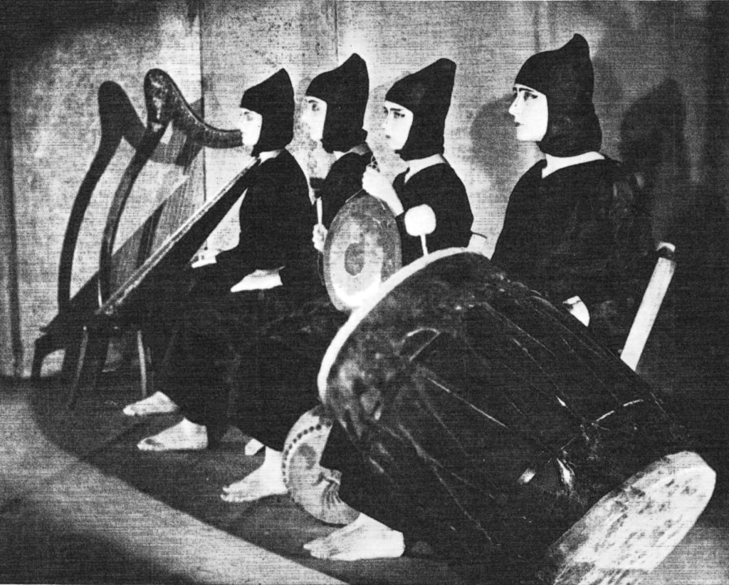 This photograph by Toyo Miatake from the book “Michio Ito: A Man and his Dances” by Helen Caldwell, Ito’s student and a participant in the production, shows the costumed musicians for “The Hawk’s Well” the William Butler Yeats “Noh” play. Caldwell describes the production: “Although “At the Hawks Well” was mounted with far greater sophistication than that of the chandelier lit drawing room advocated by Yeats, it still had no theatricality in the usual sense; the effect was poetic simplicity. Argus Bowl was a small Greek-style theatre in a hillside on the Argus estate. It seated about three hundred persons. High above the actors one saw in dim outline the jagged peaks of a sierra. All around in the dark were trees. And there was a wizard named Lewis Barrington behind the lights to give the scene the otherworld reality Yeats had sought. This new setting still called upon the spectator’s imagination, with its black night, the faint suggestion of hills, and light that seemed a part of the verse”
